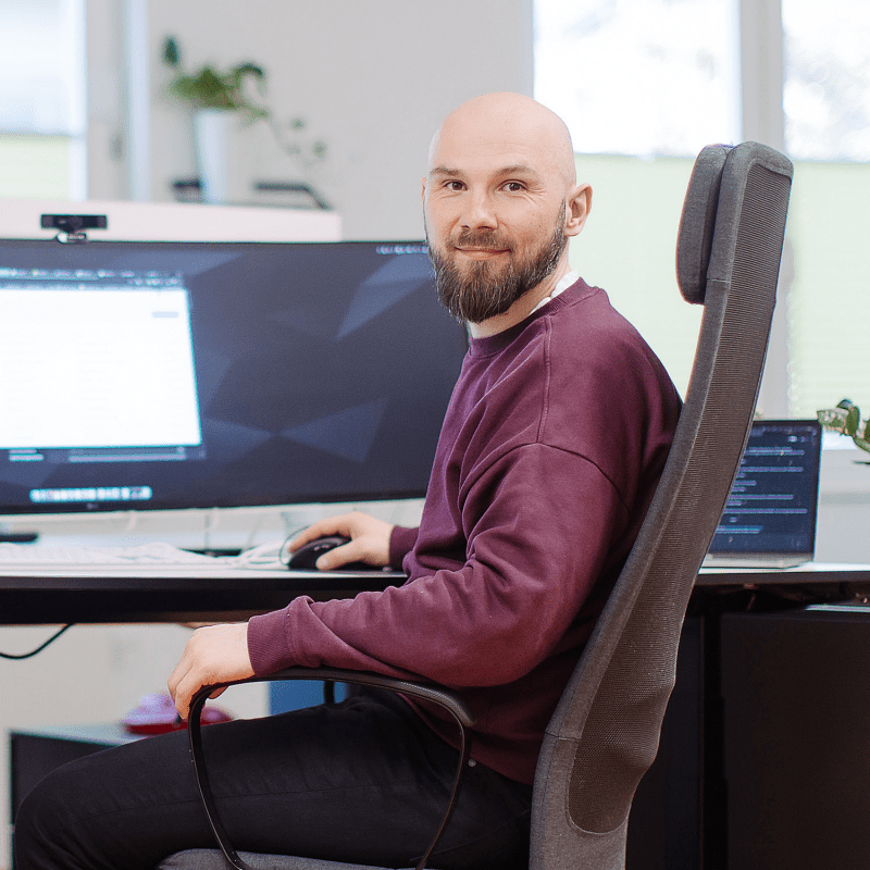 Eine Person sitzt an einem Schreibtisch vor einem gro&szlig;en Monitor, arbeitet am Computer und blickt freundlich in die Kamera.