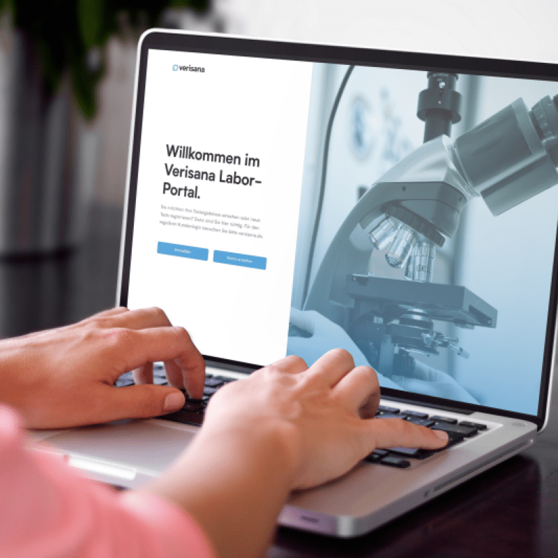 Eine Person tippt auf einem Laptop, auf dessen Bildschirm die Startseite des Verisana Labor-Portals mit Login-Optionen und einem Mikroskop zu sehen ist.
